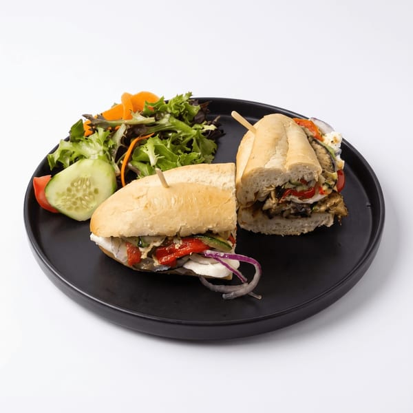 Two toasted vegetable sandwiches with roasted peppers, onions, and greens beside a fresh mixed salad on a black plate.