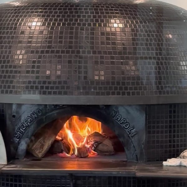 A large, dark tiled pizza oven with a roaring fire inside, ready for cooking.