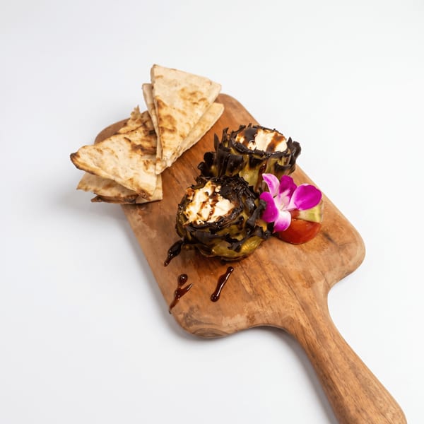 Grilled flatbread wedges accompany stuffed artichokes with creamy filling, dark sauce, tomato, and purple edible flowers on a wooden board.