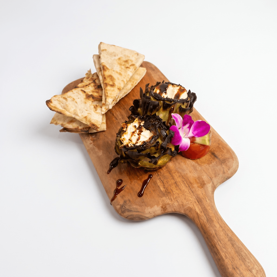 Grilled flatbread wedges accompany stuffed artichokes with creamy filling, dark sauce, tomato, and purple edible flowers on a wooden board.