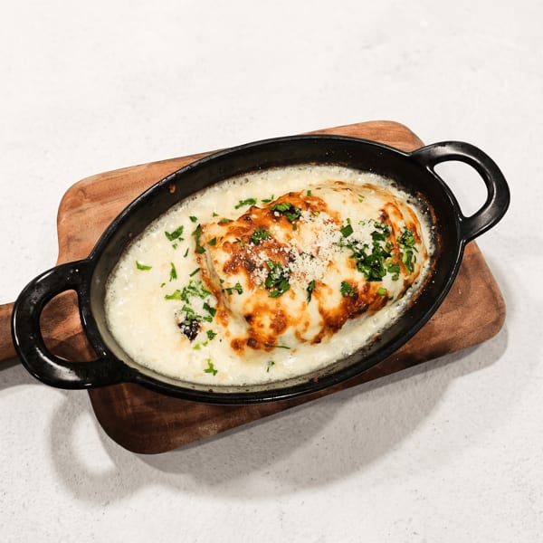 Baked cheesy chicken topped with herbs in a black oval skillet on a wooden trivet.