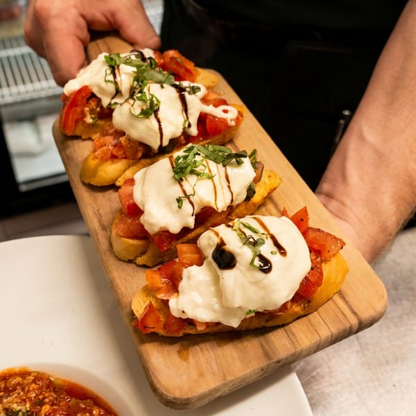 A chef's hands present a wooden board with three bruschetta topped with tomatoes, mozzarella, and basil.