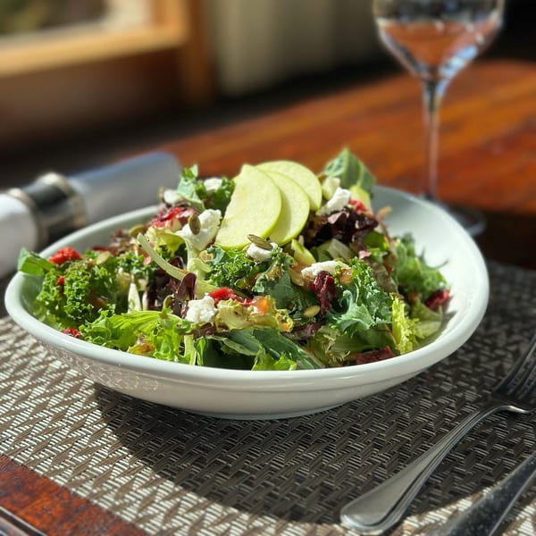 Fresh salad with mixed greens, apple slices, goat cheese, and pumpkin seeds on a table.